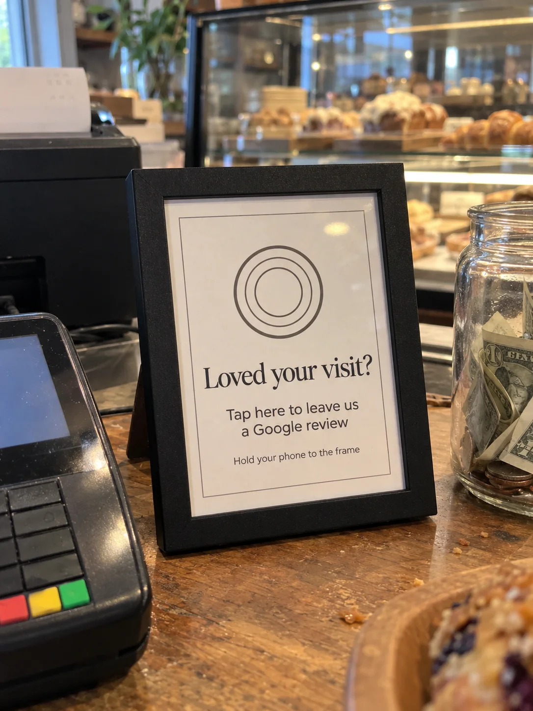 The Black TapIt Frame on its stand on a café counter beside a card reader and a tip jar